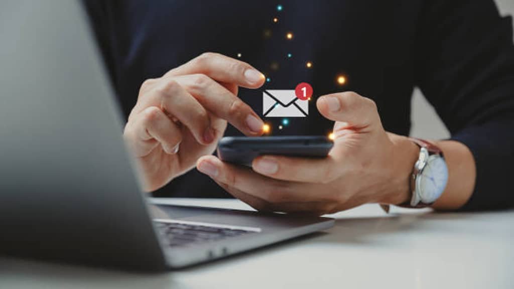 A close-up view of a person holding a smartphone with a new email notification symbol glowing, while working on a laptop, representing SMS marketing communication.