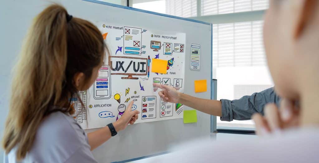 A woman points at a whiteboard adorned with various sticky notes, highlighting key ideas during a collaborative meeting